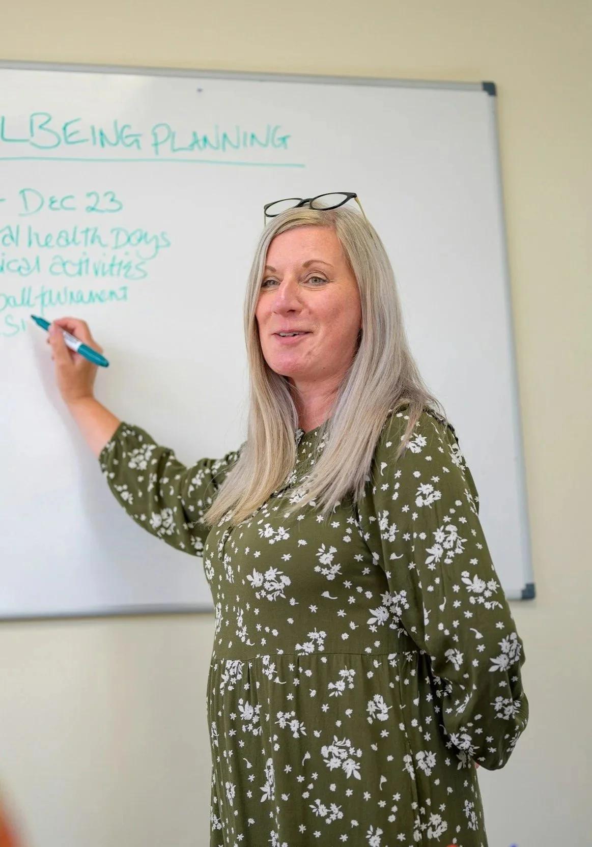 A woman with long blond hair, glasses on her head, wearing a green dress with white floral patterns, writing on a whiteboard.