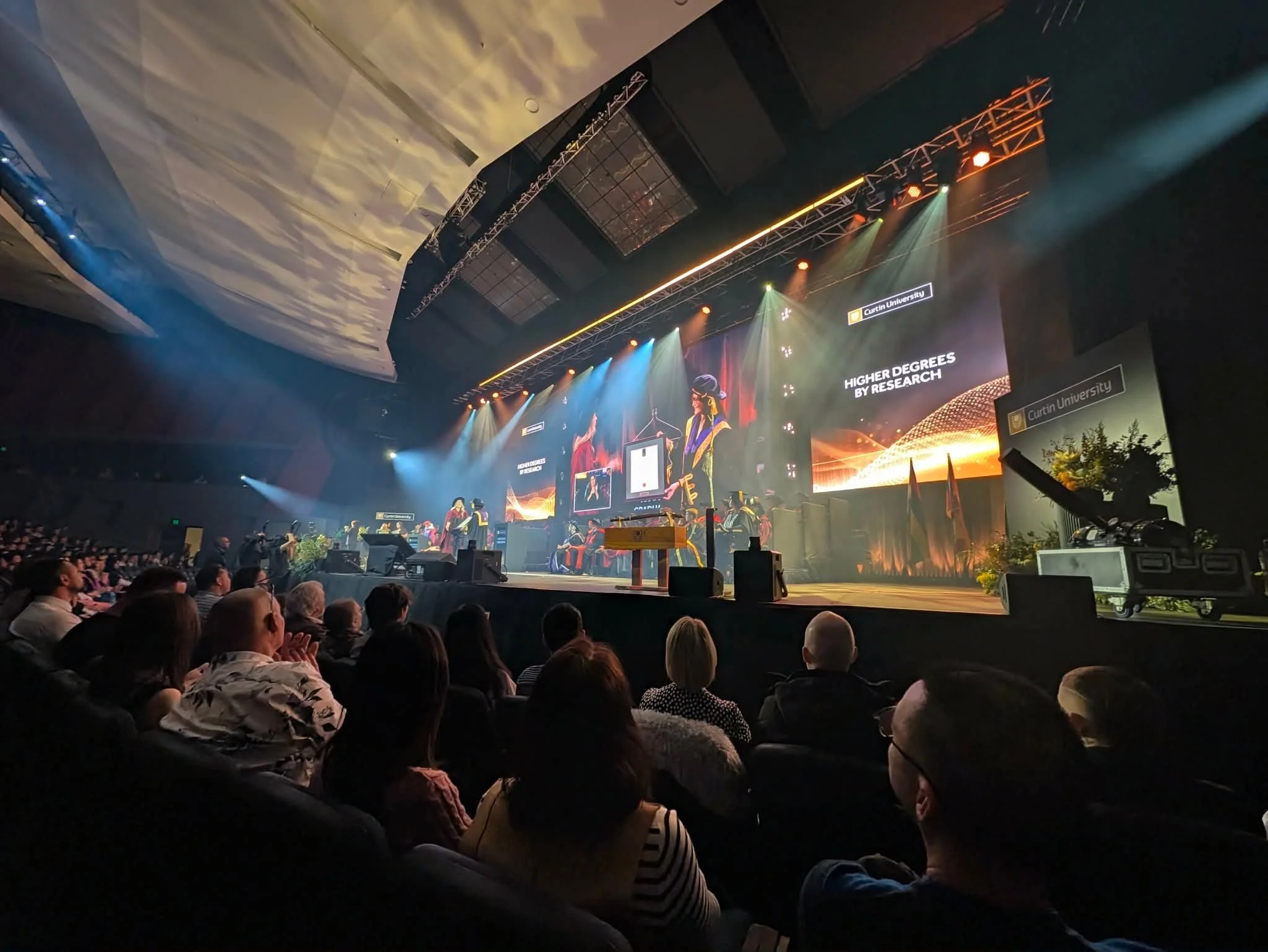 Audience seated in a dark auditorium watching a ceremony with a large screen displaying graduates and the phrase 'Higher Degrees by Research'.