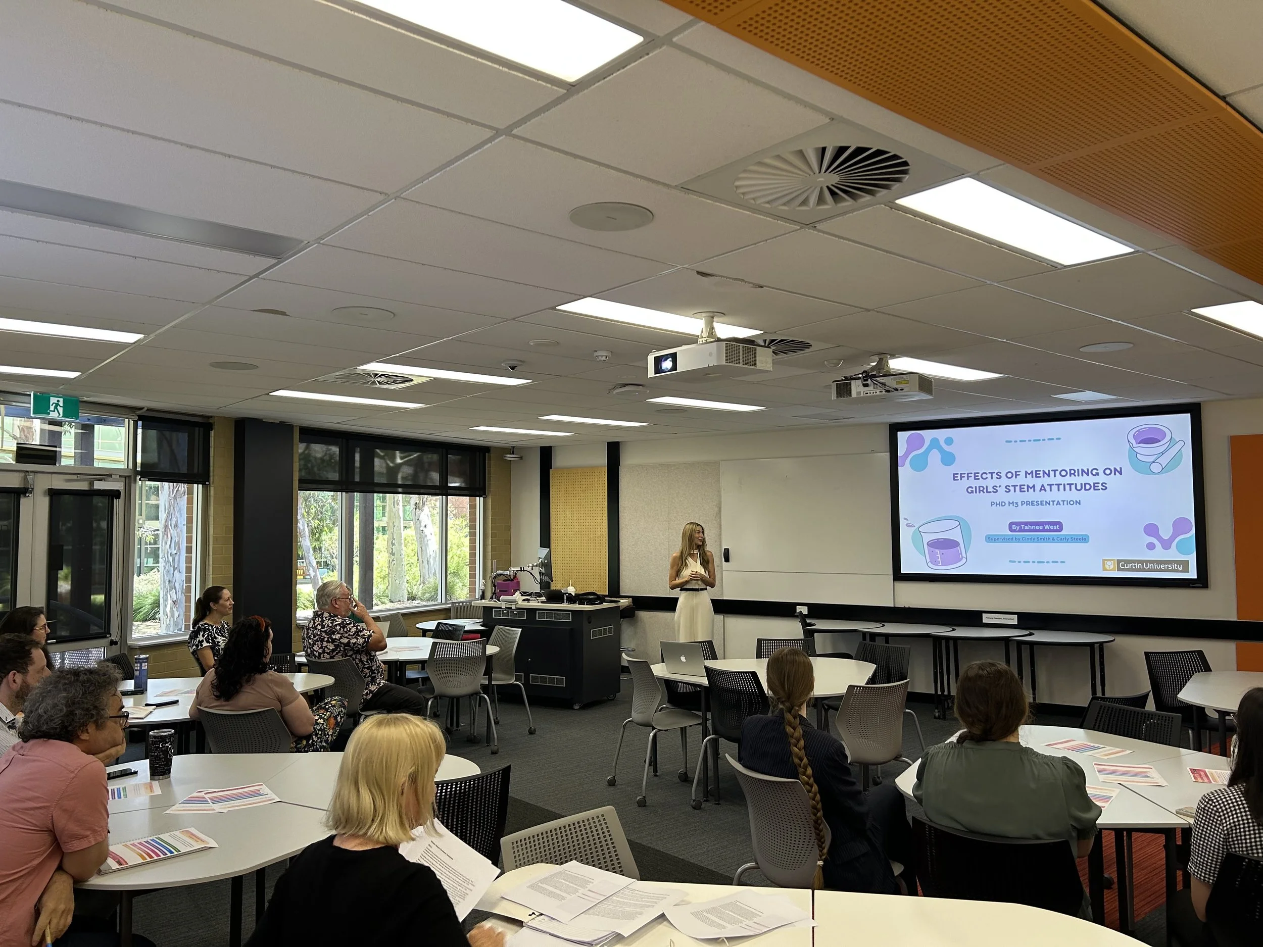 A classroom with a presentation slide displayed on a large screen. The slide reads 'Effects of Mentoring on Girls' STEM Attitudes' and includes logos from Curtin University. Several participants are seated at round tables, listening to a woman standing near the front of the room.