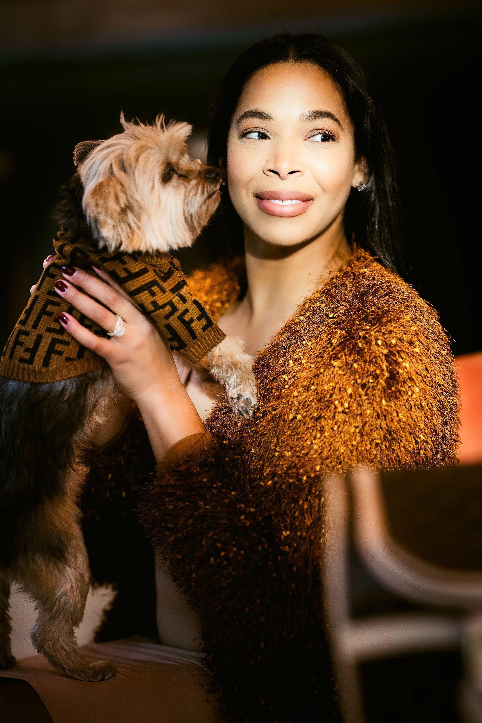 A woman holding a small dog that is licking her face in an indoor setting.