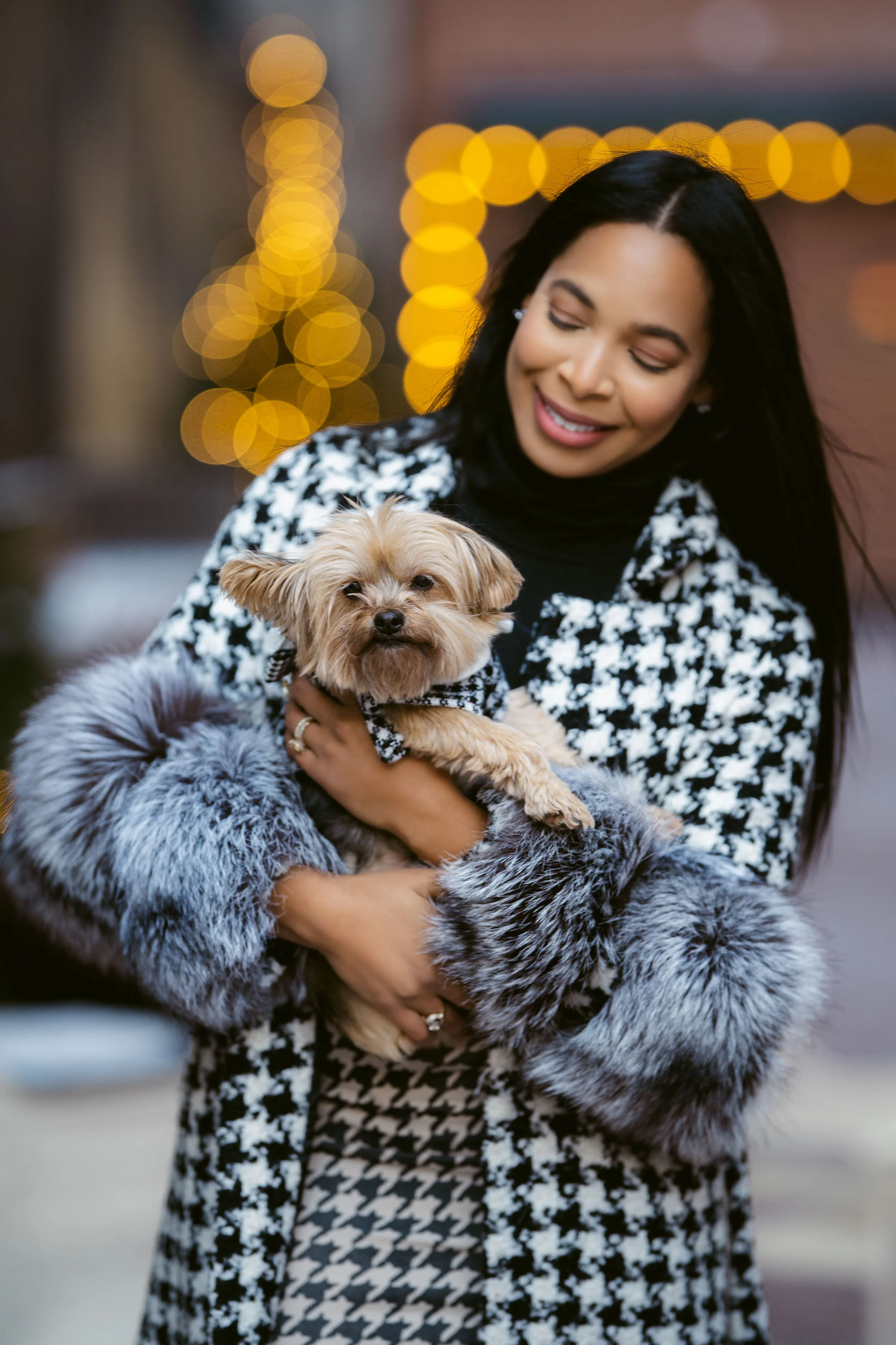 A woman smiles while holding a small dog with light brown fur. The woman is wearing a black and white patterned coat with faux fur sleeves, and the background features warm yellow bokeh lights.