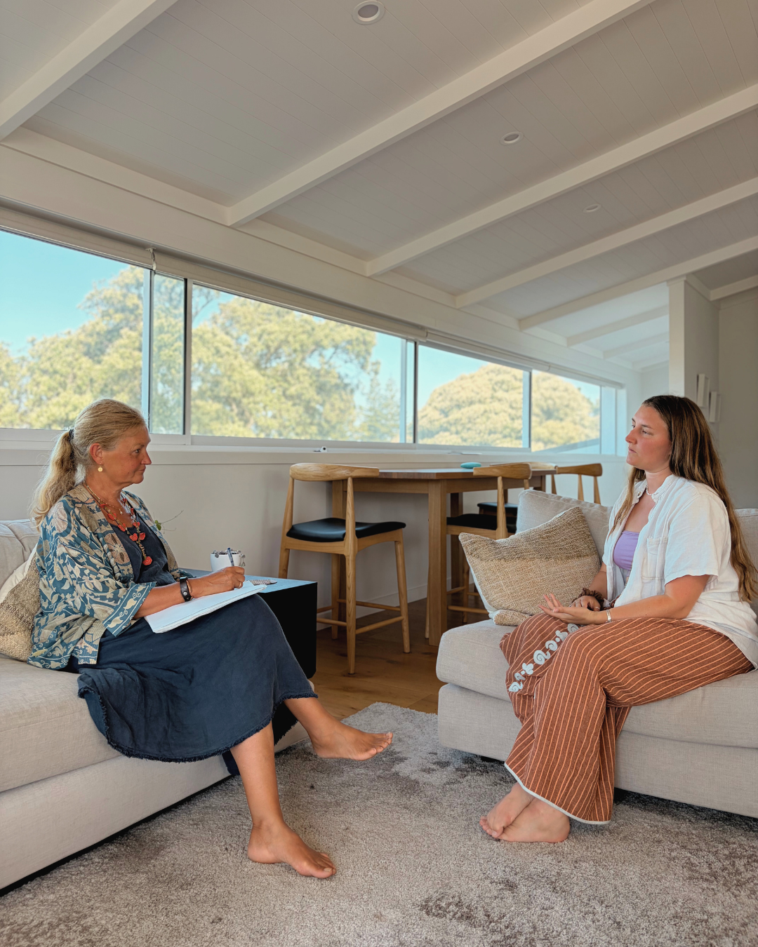 Two women having a conversation in a bright living room, sitting on separate sofas. One woman is older, with gray hair, wearing a blue dress and patterned shawl, taking notes. The younger woman has long brown hair, wearing a white shirt and striped pants, sitting crossed-legged. Large windows with a view of trees are in the background.