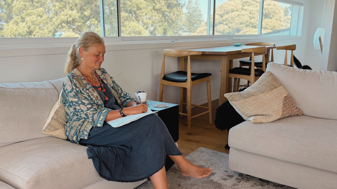 A woman with gray hair sitting on a beige sofa writing in a notebook, with a cup of tea or coffee beside her, in a bright living room with large windows, a wooden dining table with chairs, and a beige cushion on the sofa.