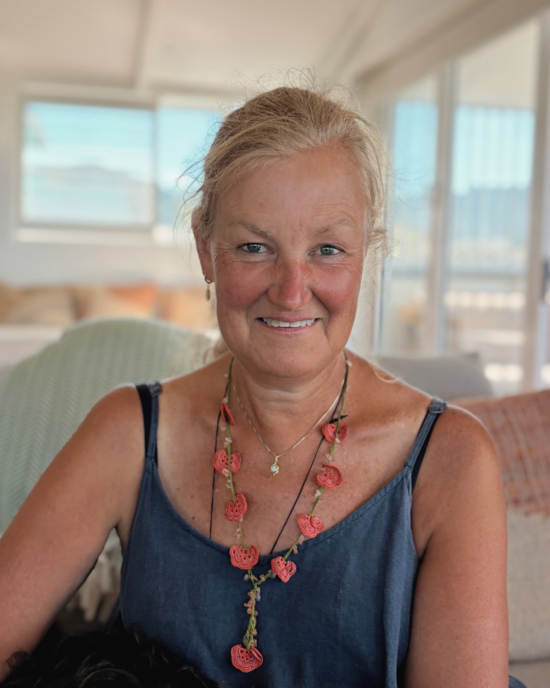 A smiling woman with blonde hair tied back, wearing a blue sleeveless top and colorful beaded necklaces, sitting in a bright room with large windows in the background.