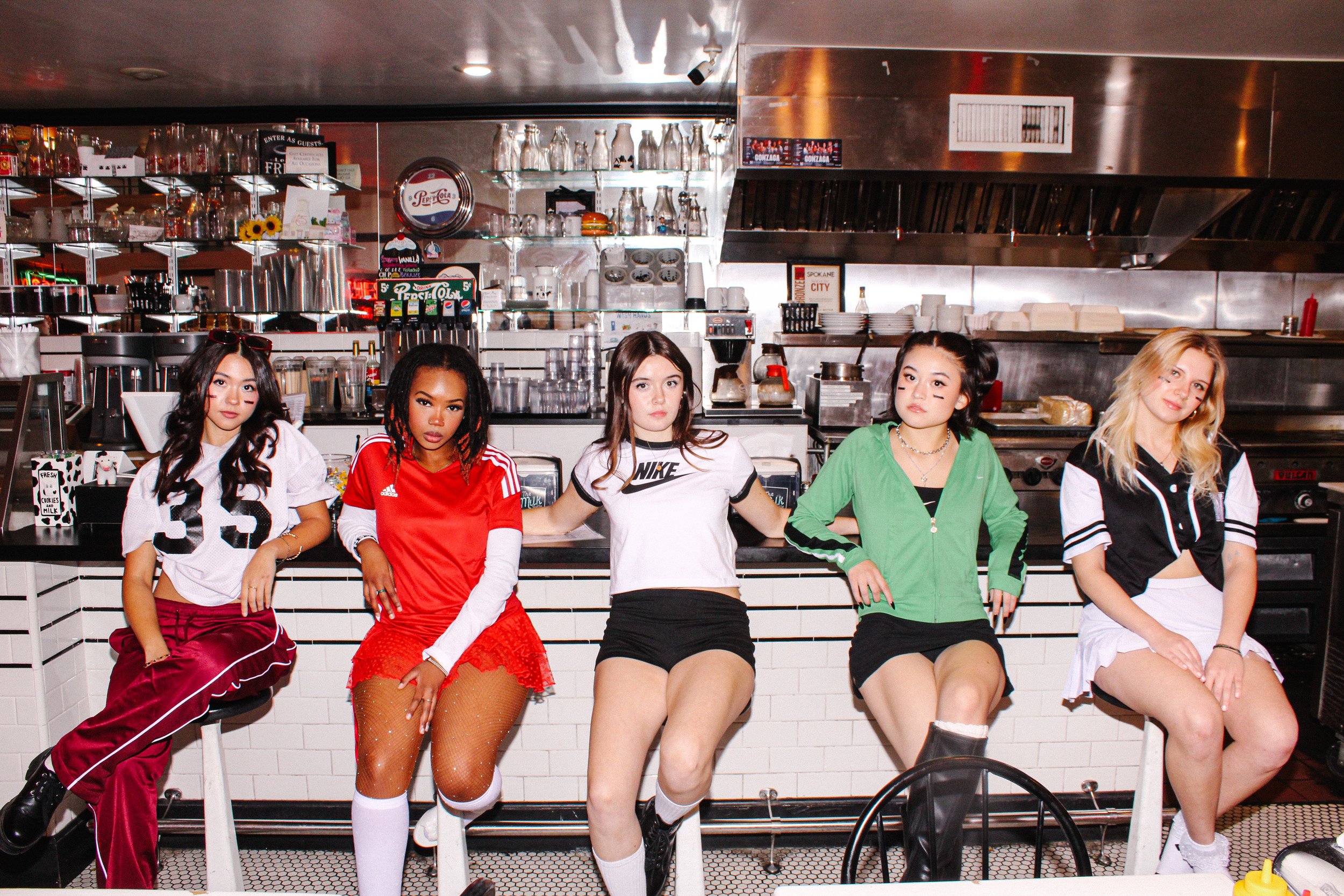 Five girls sitting on bar stools in a cafe or restaurant, dressed in sporty, casual outfits. The background includes shelves with glasses, bottles, and kitchen equipment.