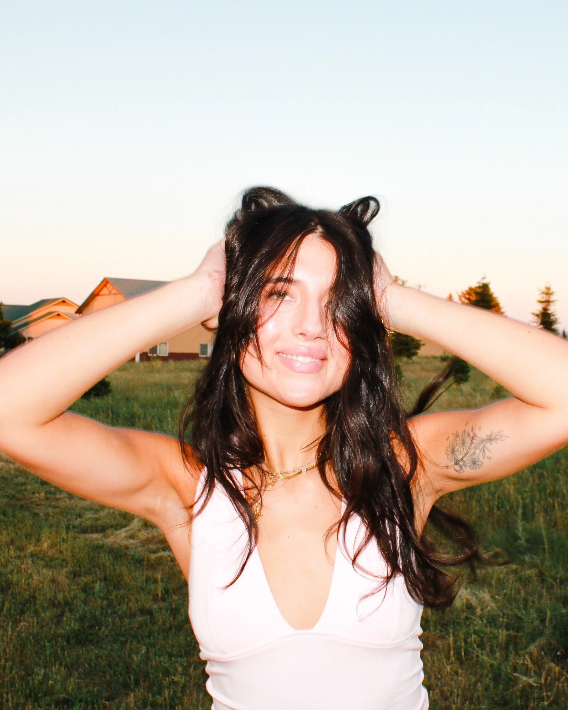 Young woman with dark hair and a tattoo on her left arm outdoors during sunset, smiling with hands on her head.
