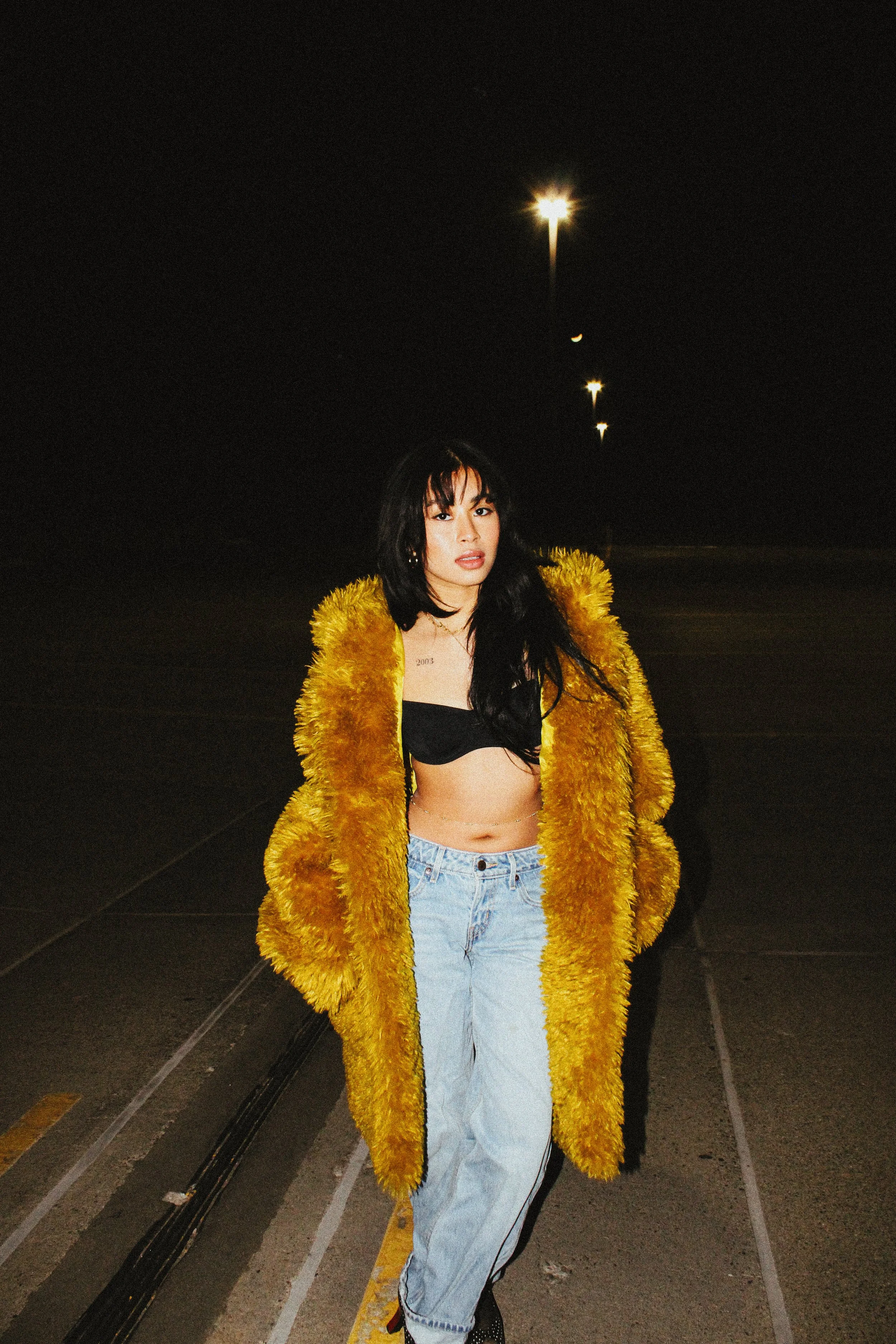 A woman walking in a dark street at night, wearing a large yellow fur coat, black crop top, and light blue jeans.