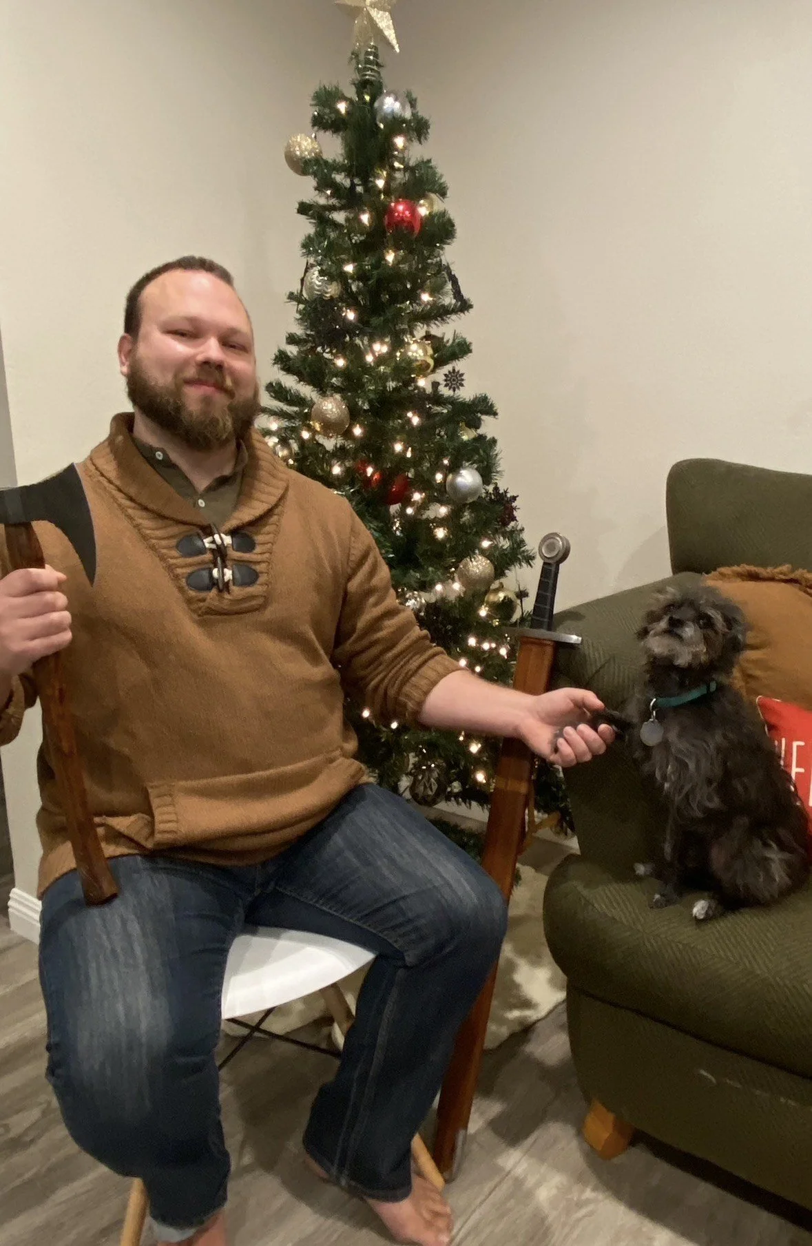 A man sitting on a chair holding a small axe, sitting in front of a Christmas tree, and petting a small black dog sitting on the arm of a green sofa.