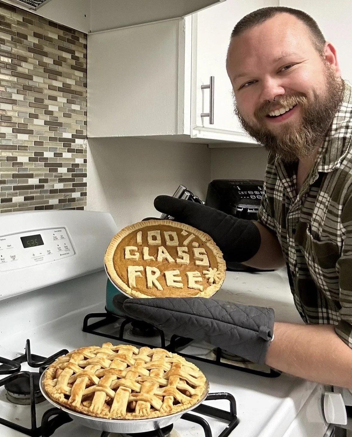 A man with a beard and a checked shirt smiling in a kitchen, holding an apple pie with the words '100% Gluten Free' spelled out in pie crust on top. There is another freshly baked pie with a lattice crust on the stove in front of him.