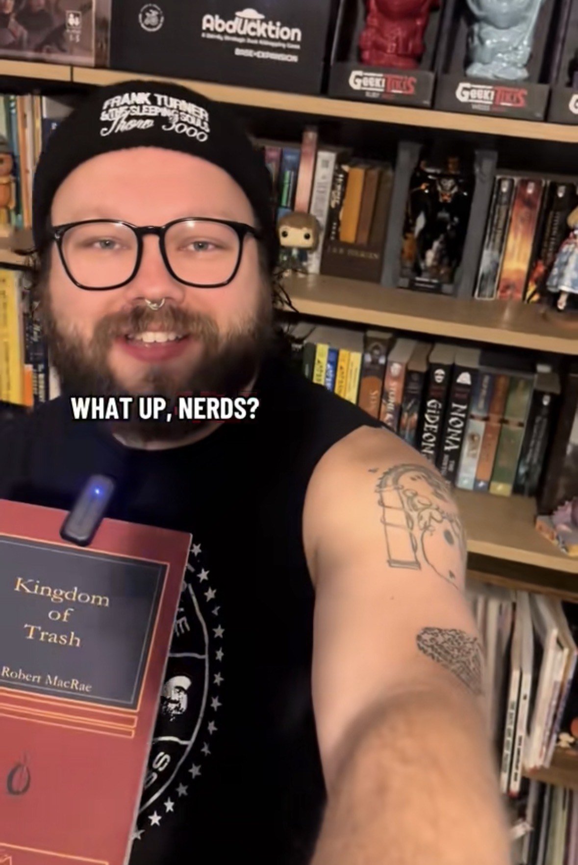 A man with glasses and a septum piercing, wearing a black beanie and sleeveless shirt, taking a selfie in front of bookshelves filled with books, figurines, and collectibles. Text overlay says, 'WHAT UP, NERDS?'.