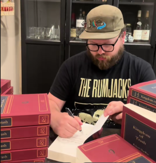 A man with glasses and a beard wearing a beige cap, black T-shirt, and sitting at a table, signing books surrounded by stacks of red and black books titled 'Kingdom of Fash'.
