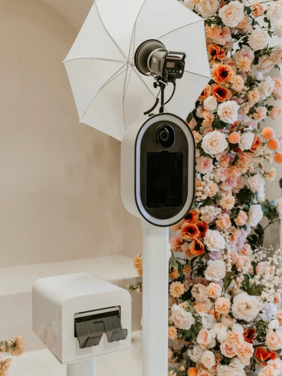 A modern  13 inch ipad pro dslr photobooth with specialized photo printer and printer stand.  scene features a floral backdrop of pink, peach, and white flowers, against a beige wall.