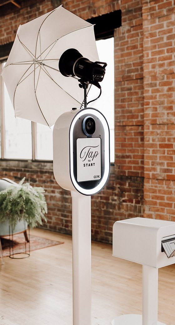 A photo booth with a large screen that says "Tap START," a dslr camera, a studio flash setup, and a white brick-wall background in a modern indoor space.