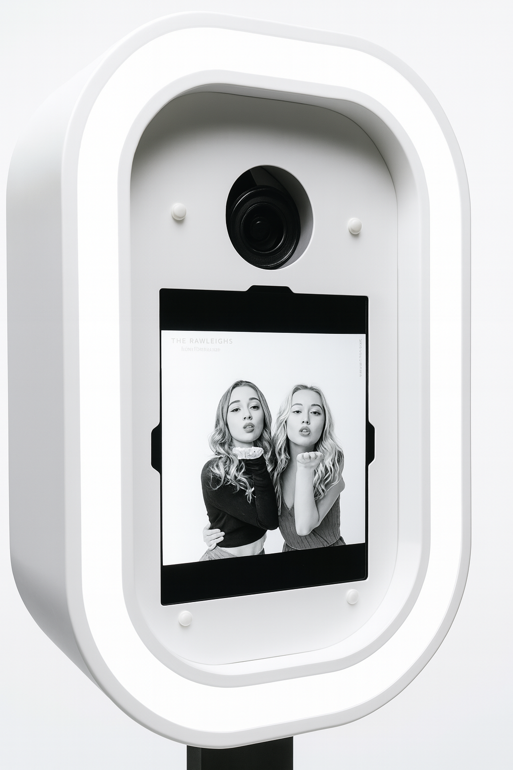 A black and white photo booth captures two young women with long, wavy hair making pouty faces, one holding her fist near her chin and the other with her hand on her waist, displayed on a touchscreen inside a modern photo booth.