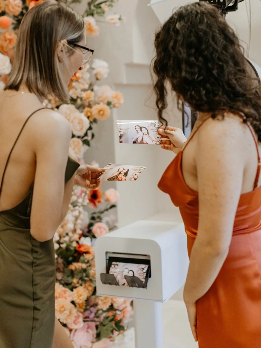 Two women in sleeveless dresses looking at photo booth pictures at a floral decorated event.