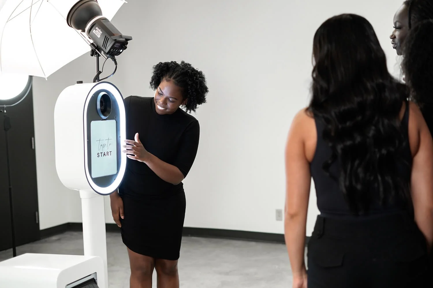 A woman is smiling and holding her hand out as she interacts with a touchscreen kiosk that says 'Tap to START.' Two women stand nearby, observing the scene, in a room with professional lighting equipment.