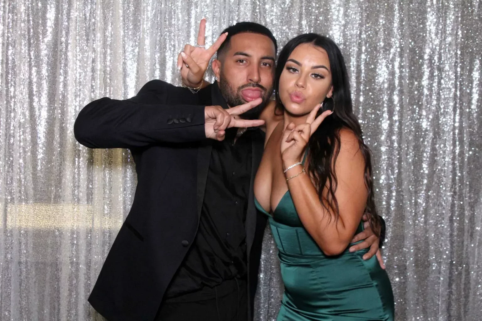 Two people posing together in front of a sparkly silver backdrop, making peace signs and playful faces. The man has short dark hair, facial hair, and is wearing a black suit. The woman has long dark hair, is wearing makeup, and is dressed in a green,