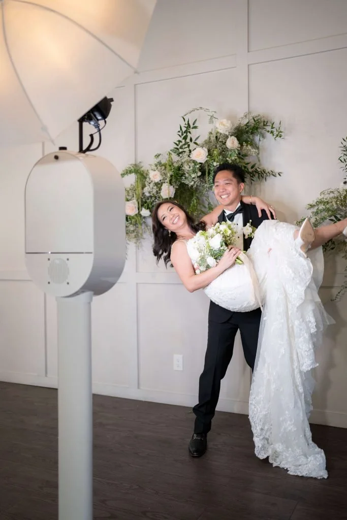 A bride and groom smile in a photo booth, with the groom lifting the bride in his arms. They are dressed in wedding attire, and the background features floral arrangements.