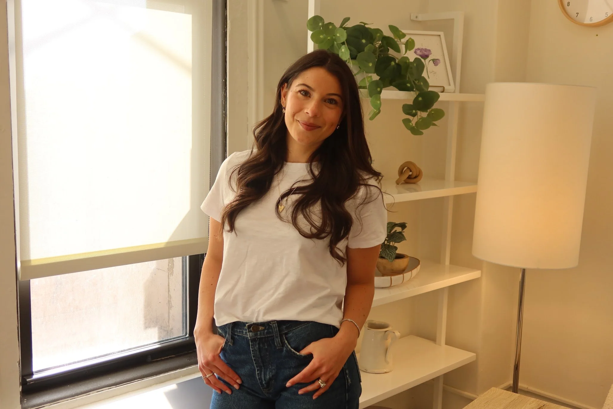 A young woman with long dark hair, wearing a white t-shirt and blue jeans, standing indoors near a window with sunlight, smiling, with a white shelf and houseplants in the background.