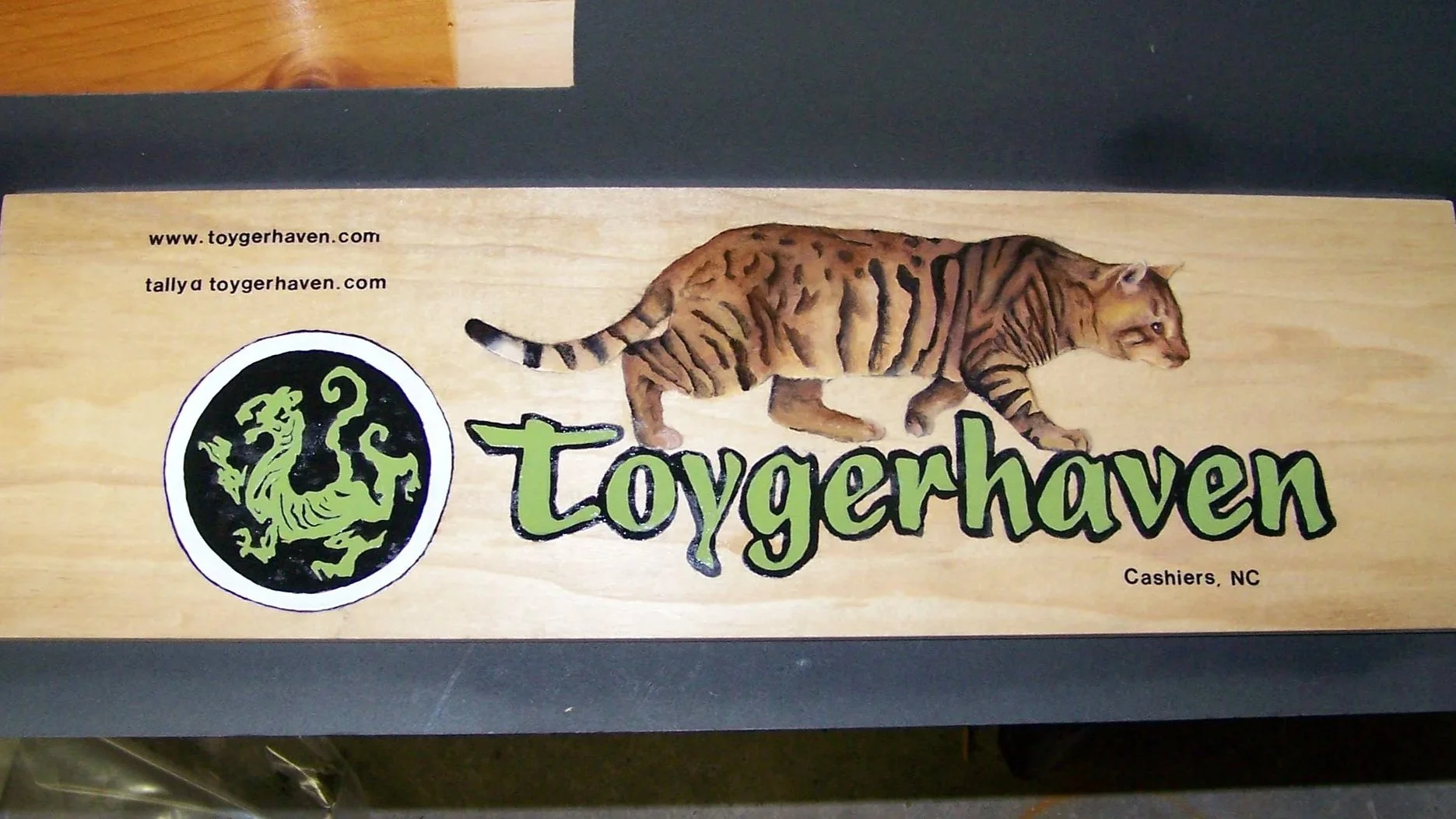 Wooden sign for Toygerhaven featuring a tiger walking to the right, a black and green circular logo with a stylized tiger face, and green text reading 'Toygerhaven' with the website address and location 'Cashiers, NC'.