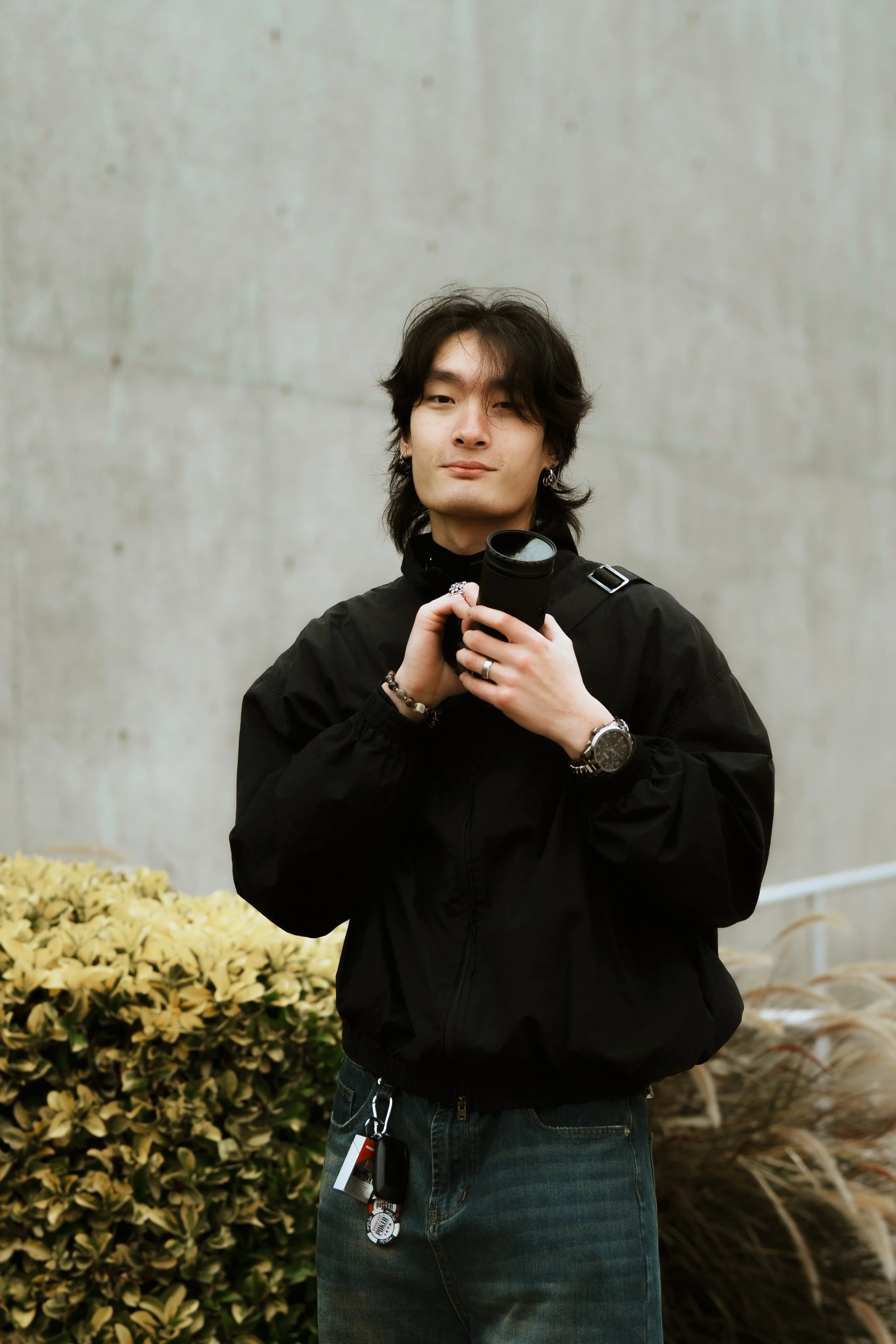 A young man with dark, wavy hair and earrings, wearing a black jacket and jeans, holding a camera, standing outdoors near a bush with yellow flowers and a concrete wall.