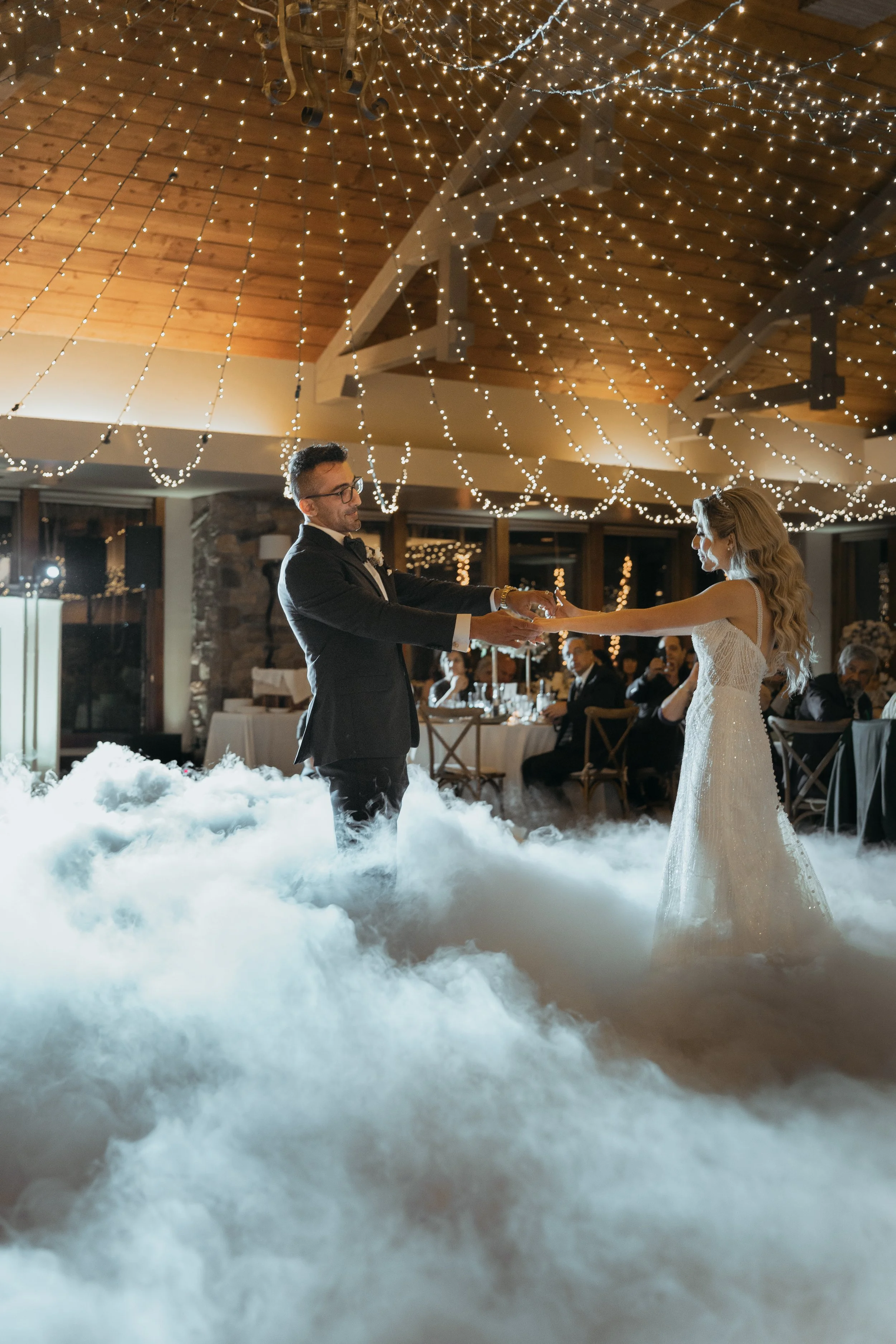 A bride and groom dancing during their wedding reception, with cloud-like fog on the floor and string lights overhead.