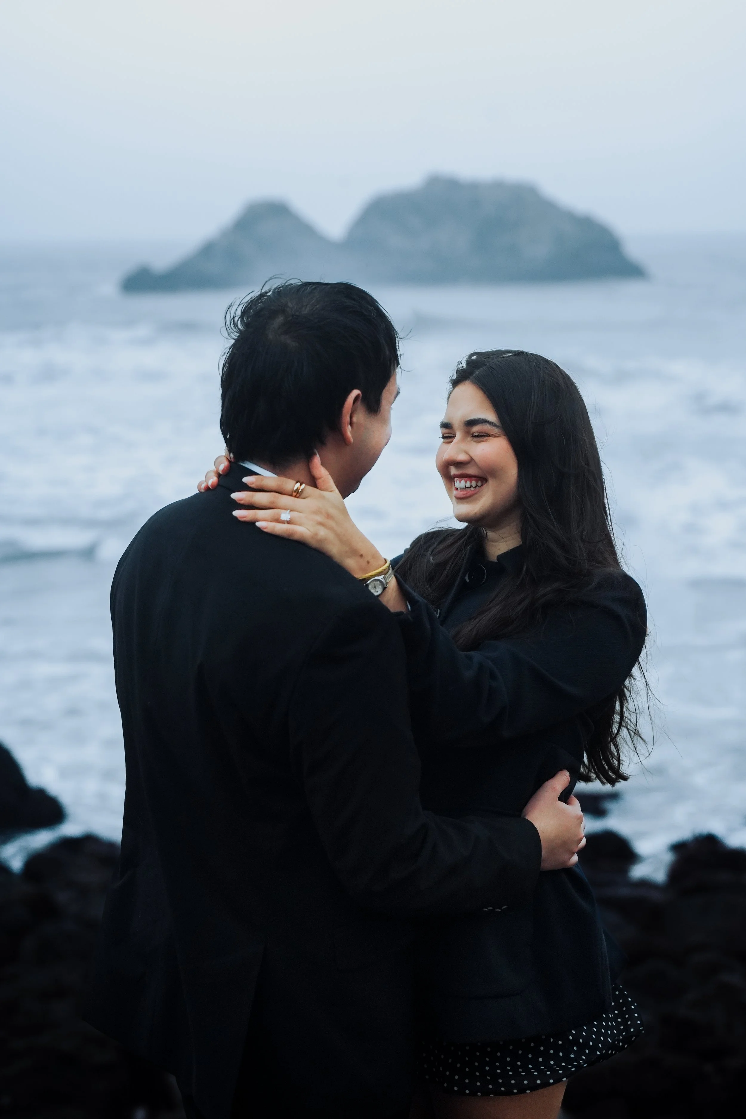 Couple embracing and smiling during a natural light engagement session by the ocean in the Bay Area, photographed by Lei Photography Collective.
