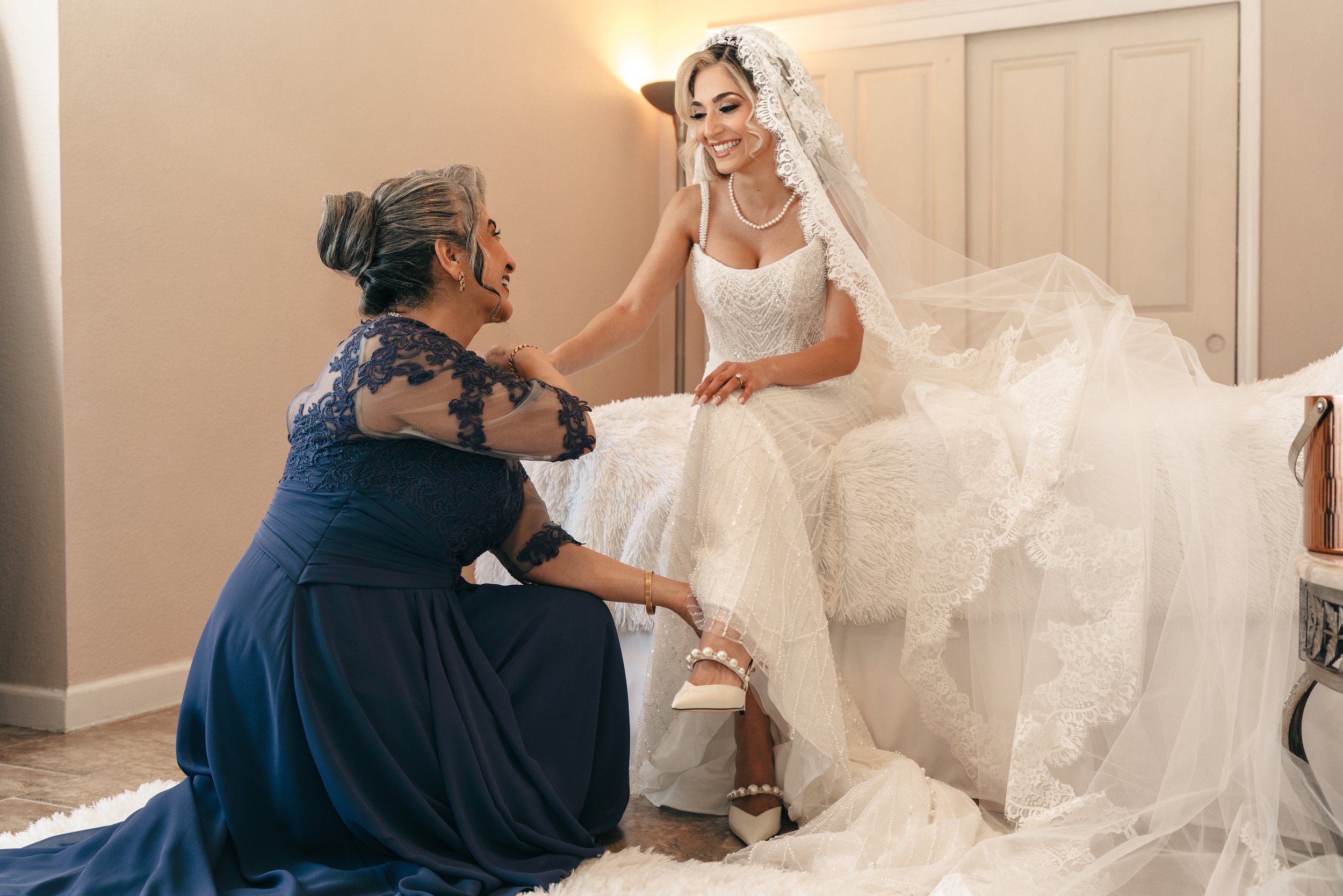 A bride in a white wedding dress is sitting on a bed, smiling, while an older woman in a navy dress kneels beside her adjusting her shoe.
