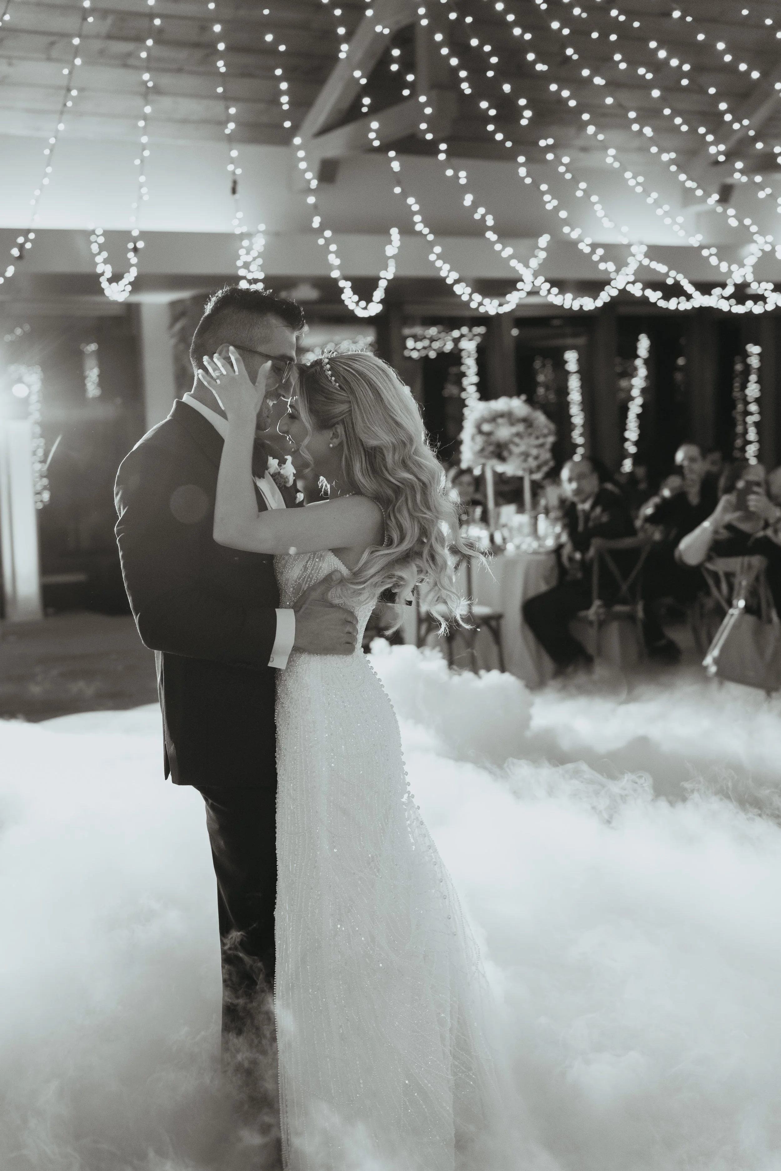 A bride and groom share a dance at their wedding reception decorated with string lights and floral centerpieces, with guests watching and taking photos.