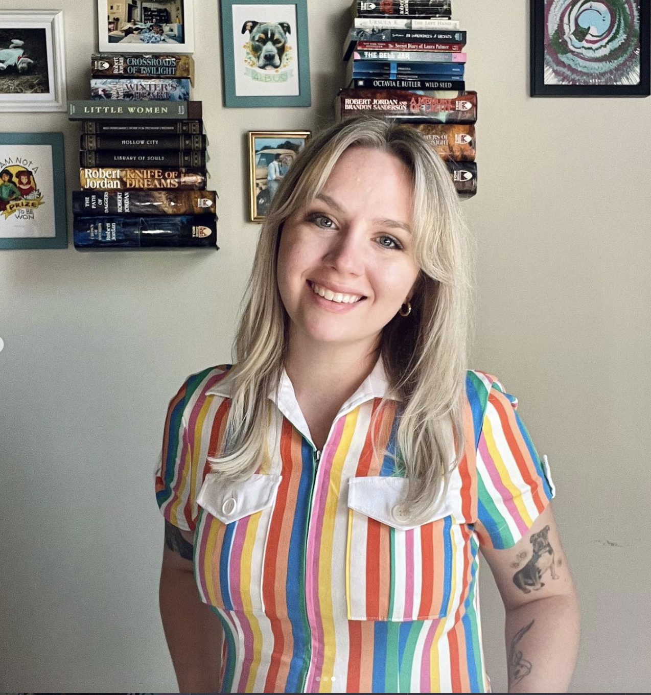 Photo of Mikelanne with shoulder length blonde hair and striped shirt smiling and standing in front of book.