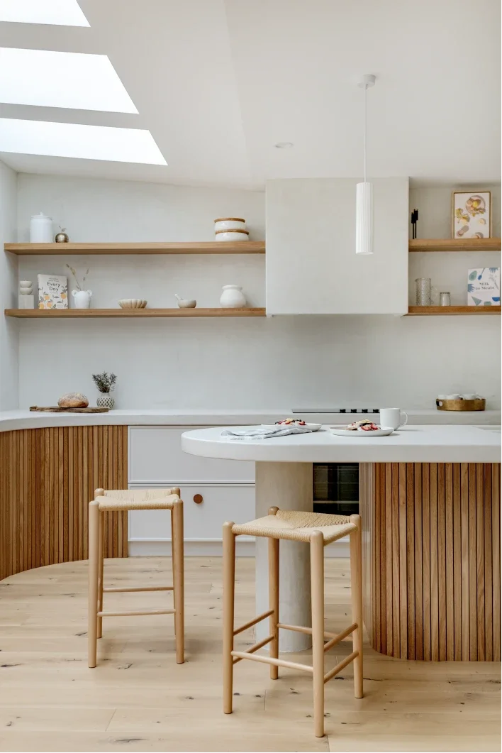Minimalist kitchen with light wood barstools, white walls, open shelves with dishes, a skylight, and a white kitchen island.