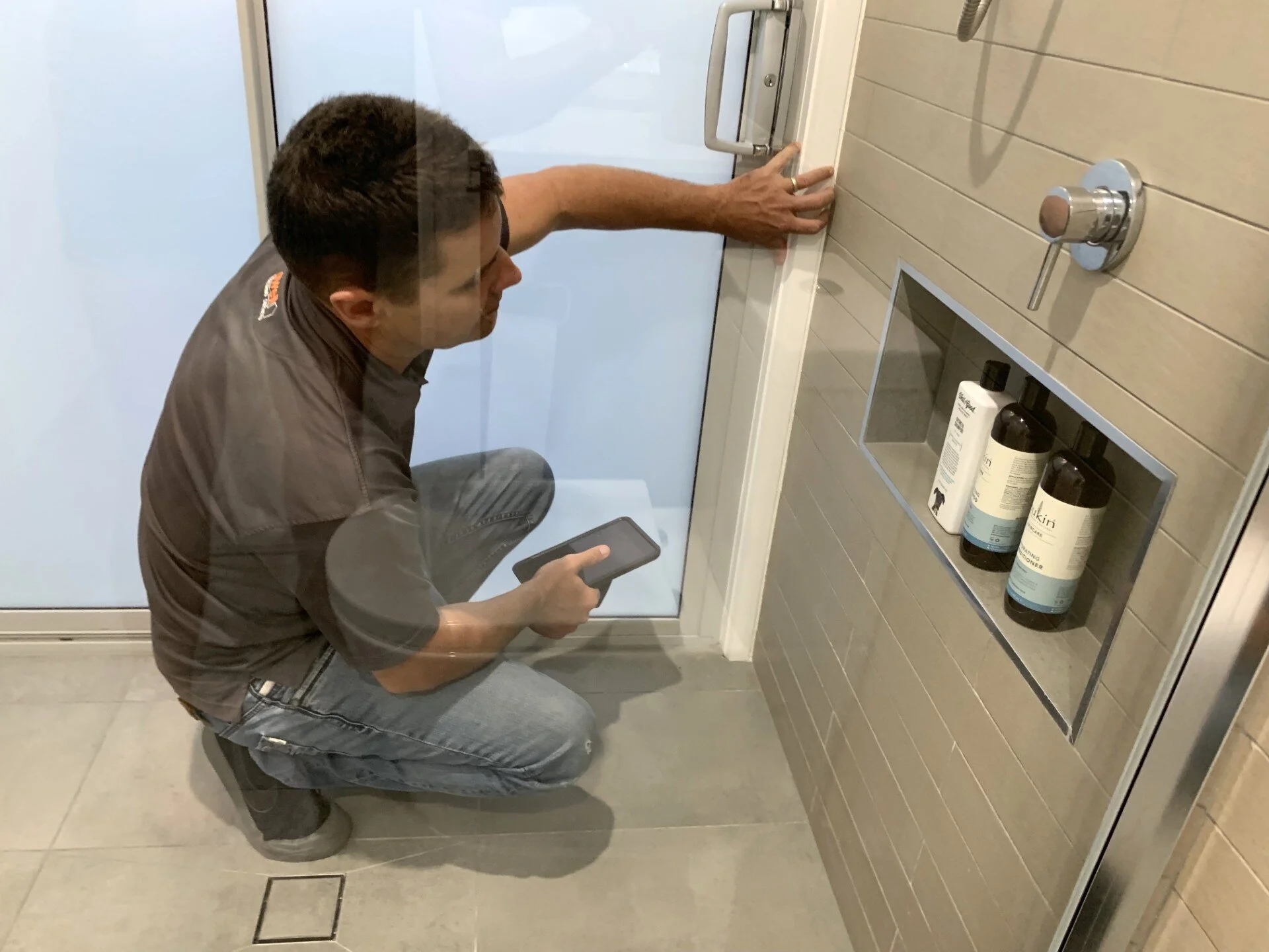 A man crouching in a shower area holding a smartphone, with his right hand touching the shower door handle. The shower wall has a built-in shelf with bottles of shampoo or body wash.