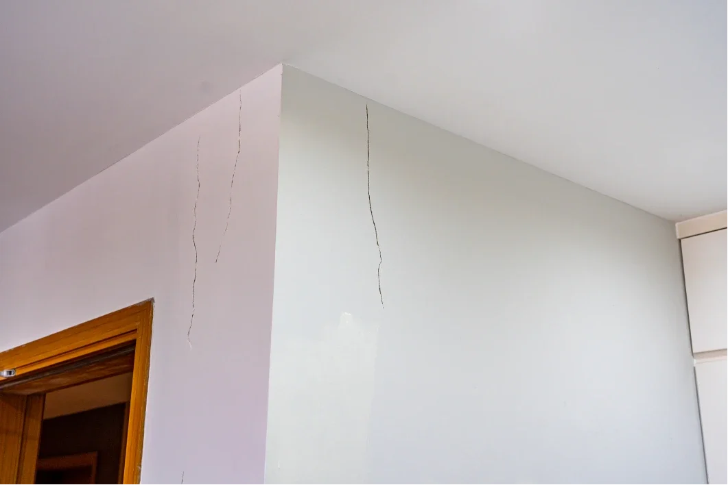 Multiple cracks in white and light-colored walls near the ceiling in a room, with a wooden door frame visible on the left.