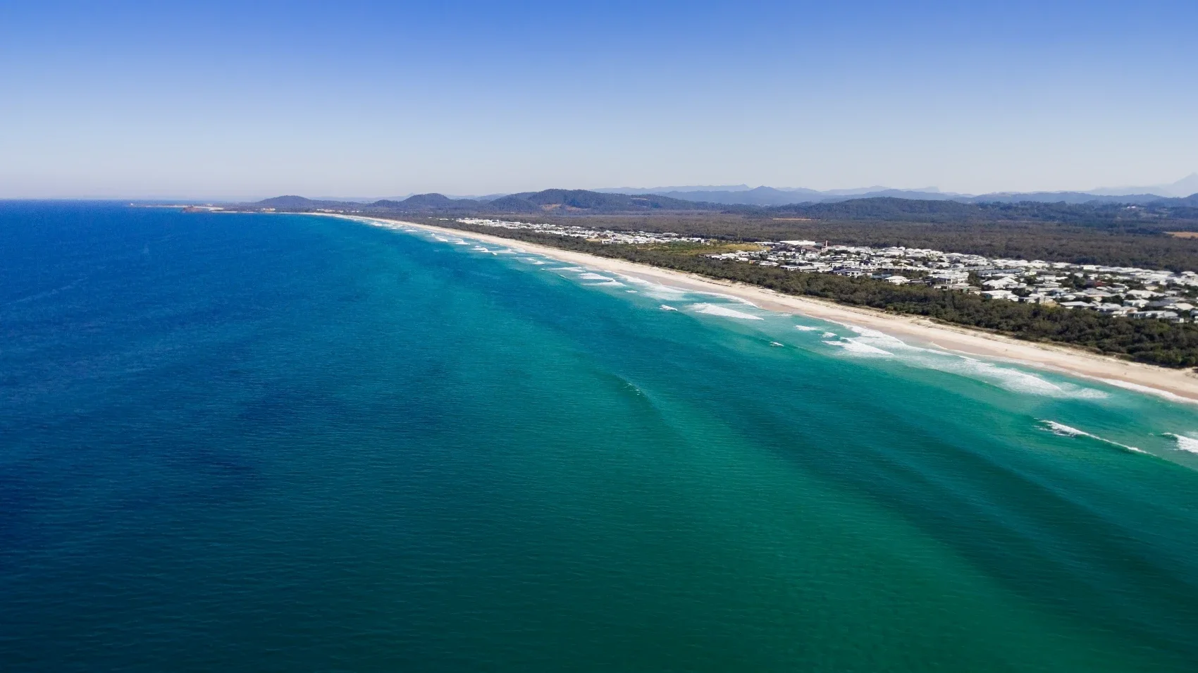 Aerial view of a beach with turquoise ocean waves and a shoreline with residential buildings, mountains in the distance under a clear blue sky.