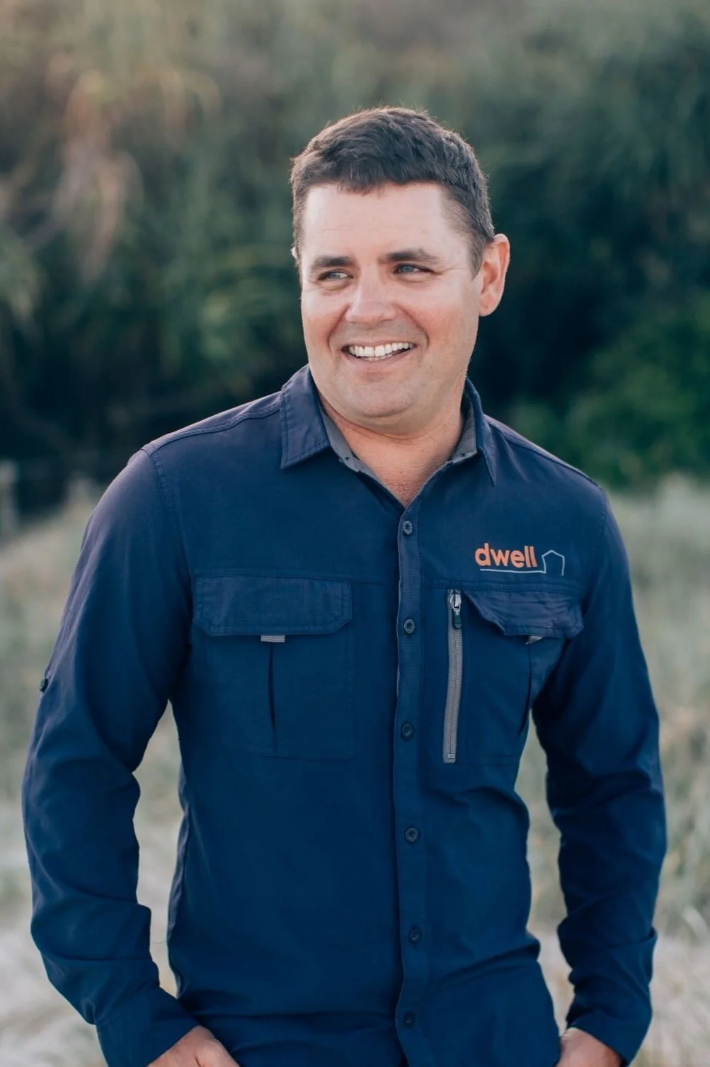 A man smiling outdoors, wearing a navy blue jacket with the word 'dwell' embroidered on it, in front of a natural background with trees and grass.