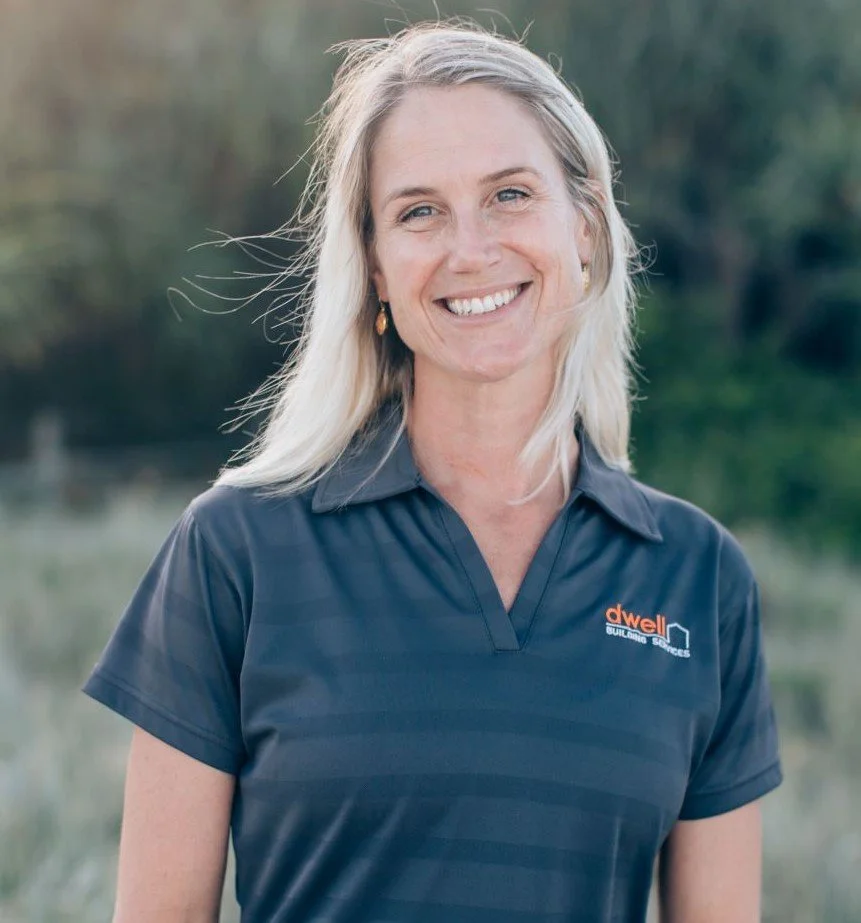A smiling blonde woman wearing a navy blue polo shirt with the Dwell Built Living Sciences logo, standing outdoors with blurred greenery and water in the background.