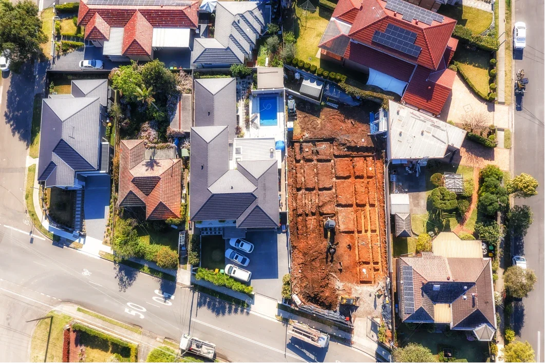 An aerial view of a residential neighborhood with houses, trees, and parked cars. The construction site in the center shows excavated soil and building foundations.
