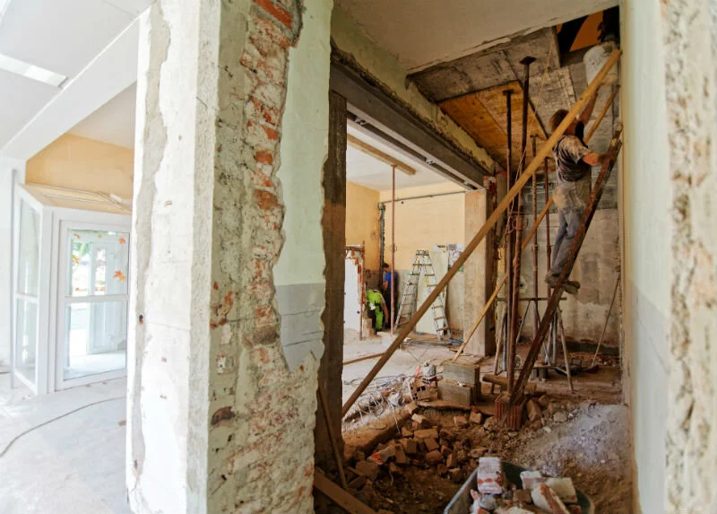 Construction workers renovating an interior space, with one worker on a ladder working on the ceiling and others in the background near scaffolding and ladders.