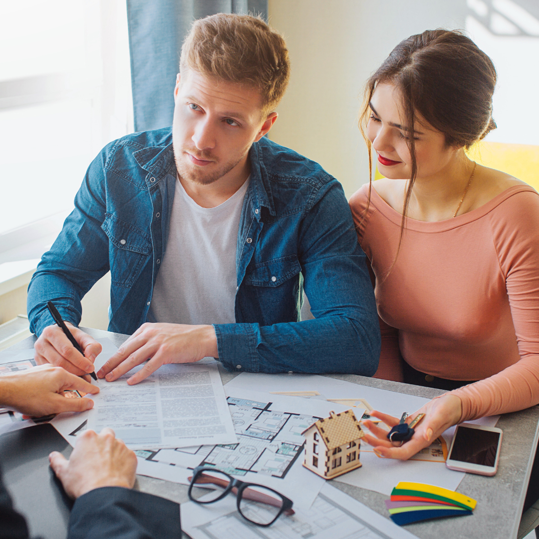 A man and woman looking at documents and a small house model on a table, discussing real estate or housing plans.