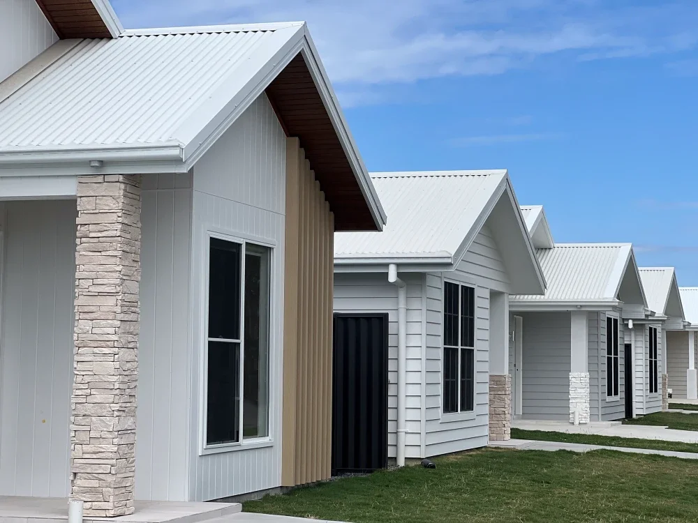 Row of modern suburban houses with white siding and metal roofs, small front yards with grass, clear blue sky overhead.