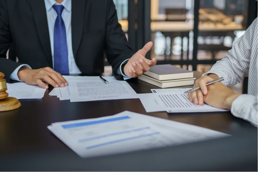 Two people in business attire sitting at a table during a meeting, with documents, notebooks, and a gavel on the table.