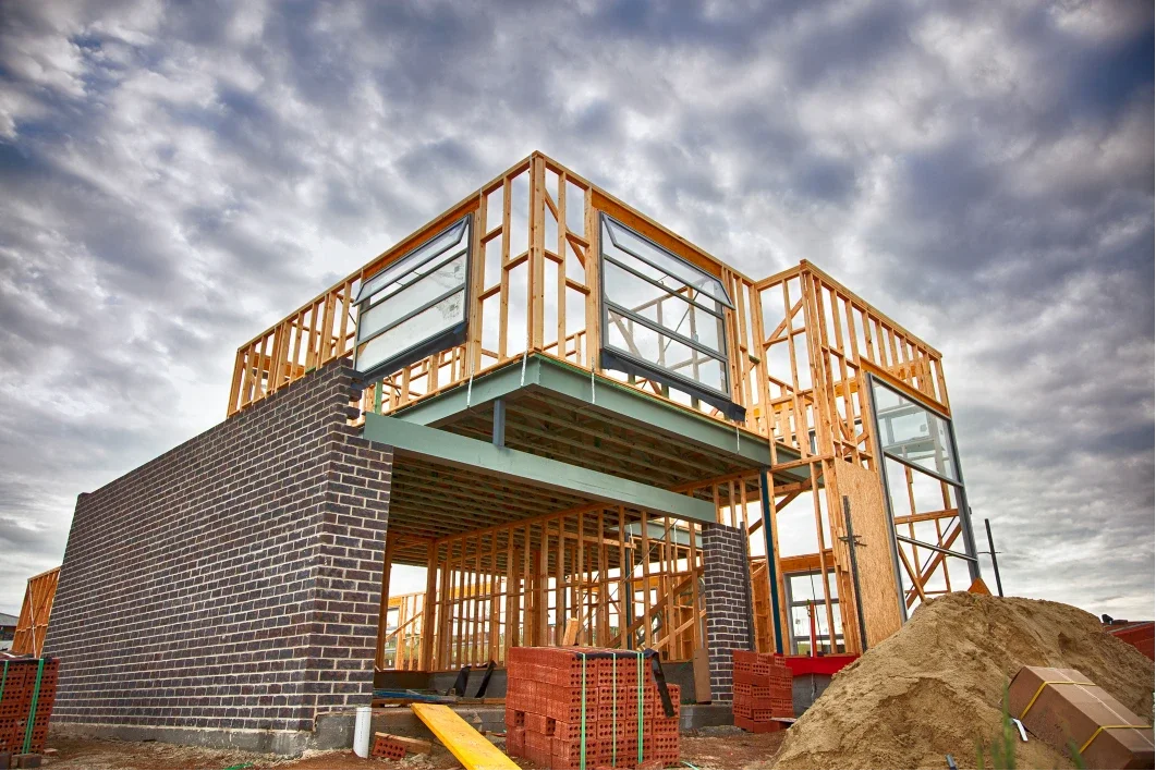 Under construction modern building with wooden framing, brick foundation, and large windows, set against a cloudy sky.