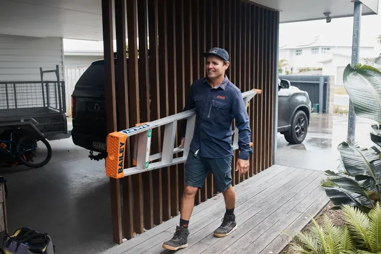 A man in work clothes carrying a ladder on his shoulder during a rainy day outside a house.