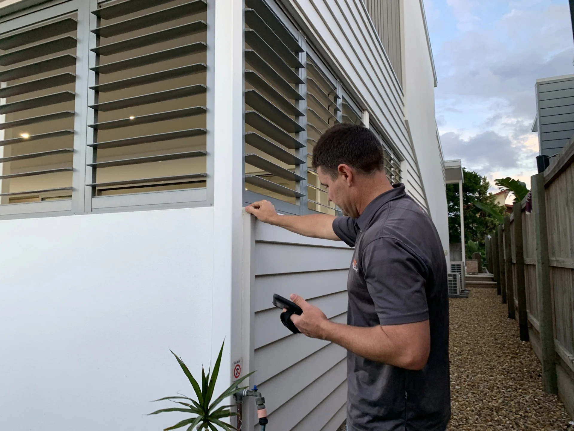 A man examining and adjusting the exterior louvered vent on a house with a phone in his hand, during the daytime.