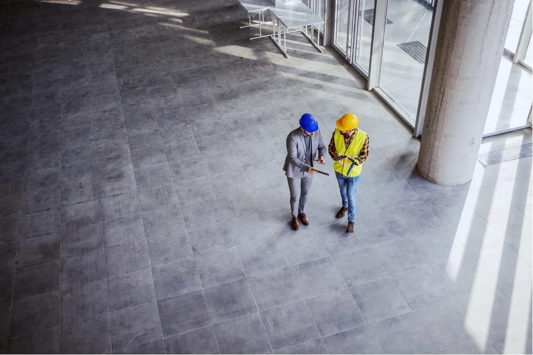 Two construction workers, one in a yellow safety vest and helmet and the other in a gray suit with a blue helmet, standing inside a large modern building with gray tiled floors and glass walls, looking at a tablet.