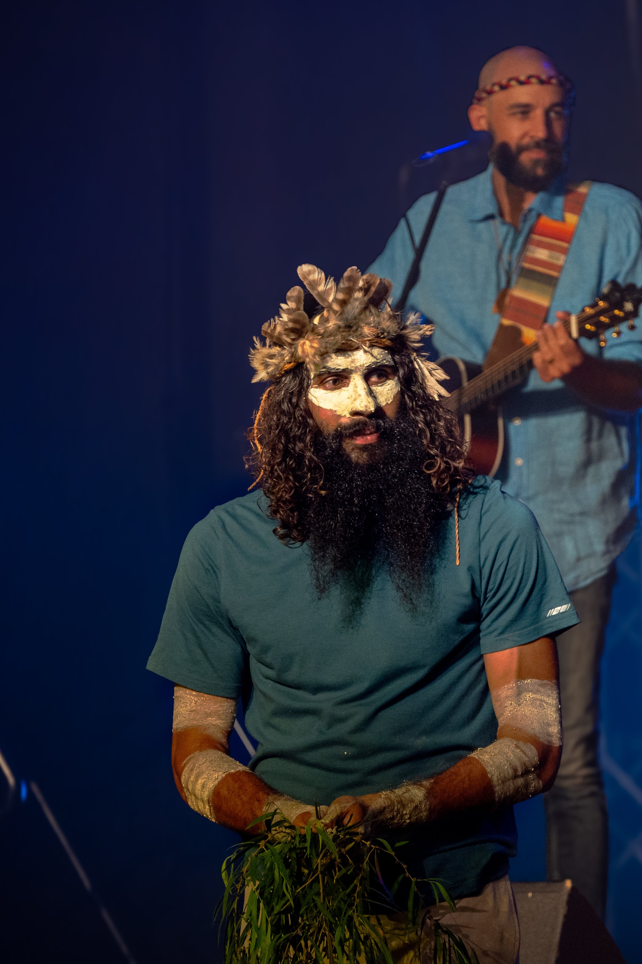 A man with face paint, a headdress made of feathers, and painted arms is sitting in front of a man playing a guitar during a performance.
