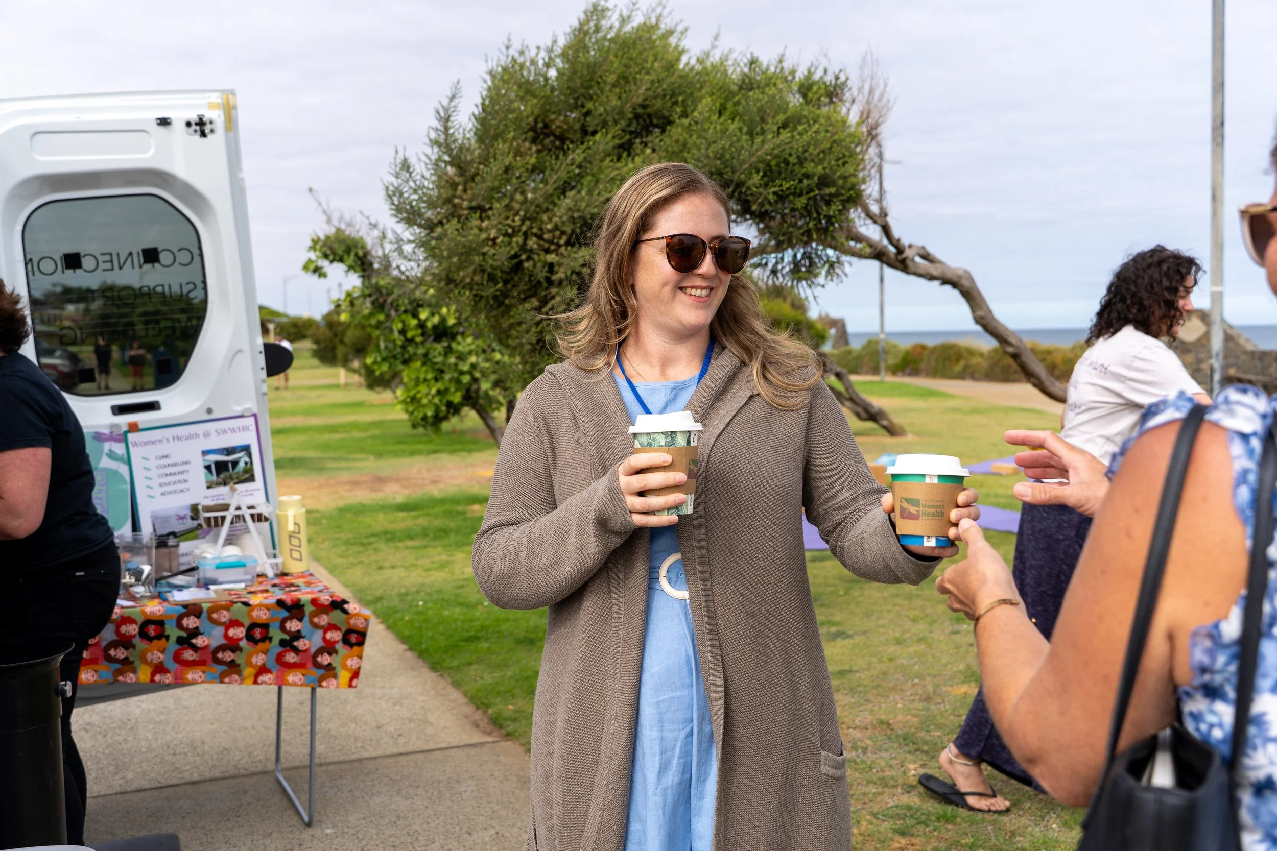 Woman with sunglasses and a light blue dress receiving a coffee cup from another woman at an outdoor event with trees and cloudy sky in the background.