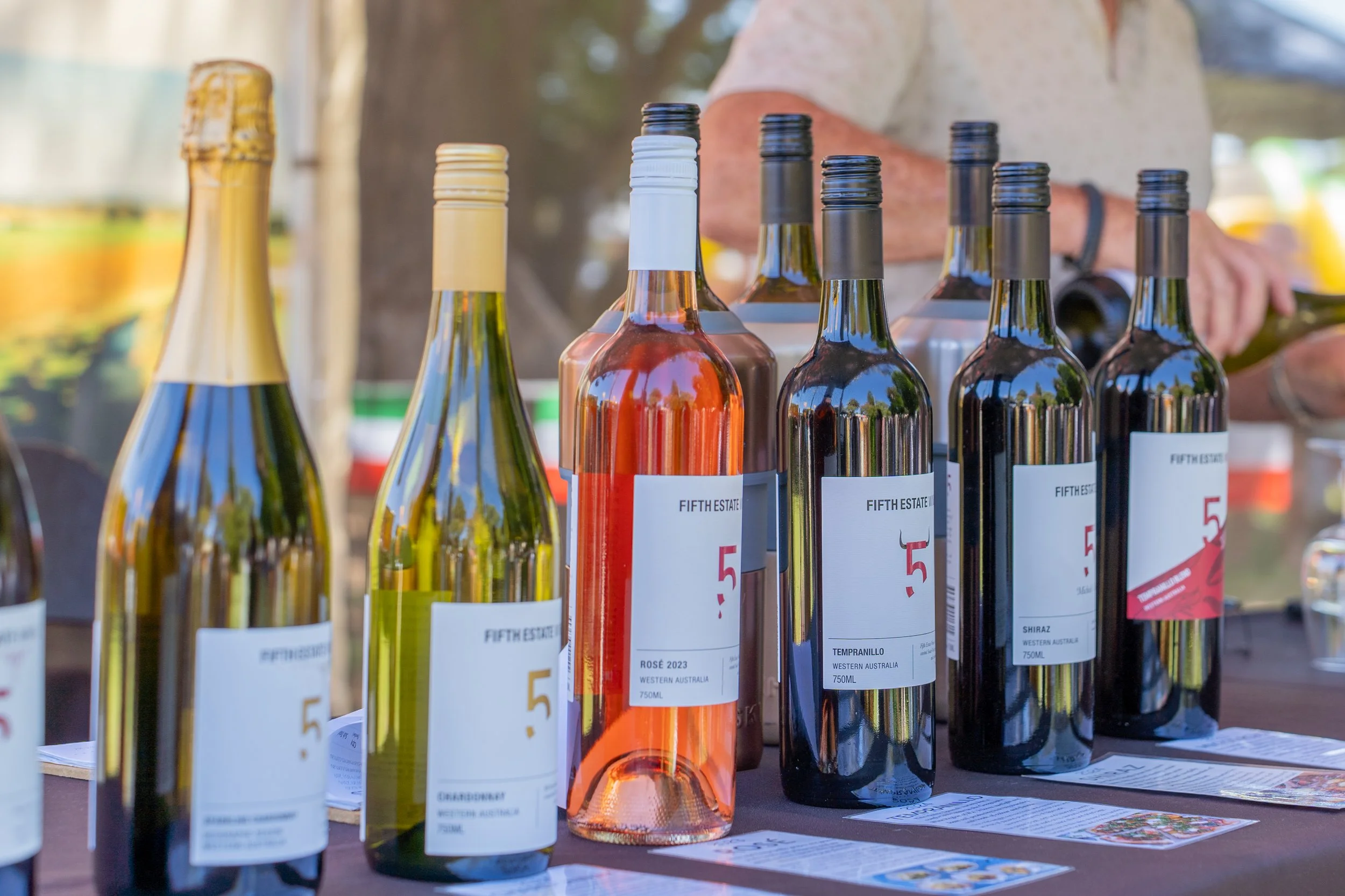 Various bottles of wine displayed on a table at an outdoor market or event, with a person in the background pouring wine.