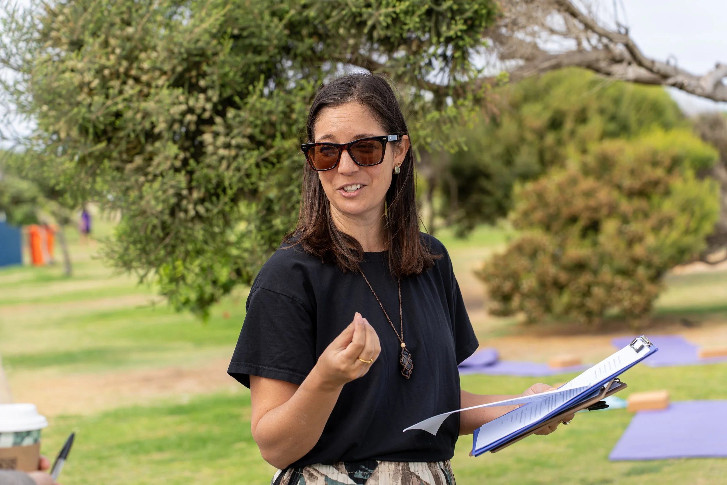A woman with shoulder-length dark hair, wearing sunglasses, a black t-shirt, and patterned pants, holding a clipboard and speaking outdoors in a park with trees and yoga mats in the background.