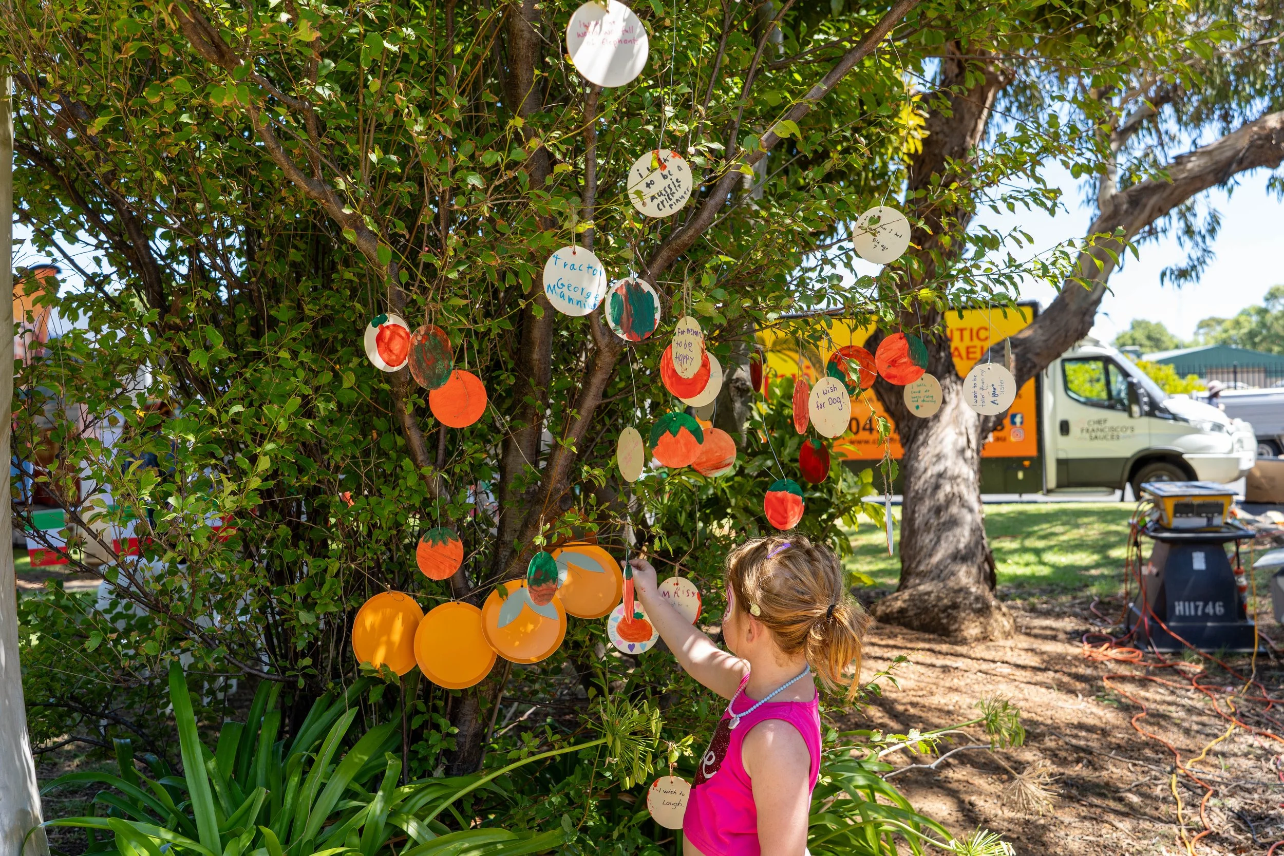 A young girl with red hair tied in a ponytail, wearing a pink top and a pearl necklace, decorating a tree with colorful paper ornaments and handwritten notes during daytime.